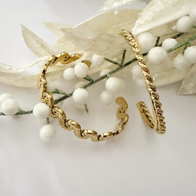 Dois brincos de argola dourados com textura e elementos ondulados junto a ramo decorativo branco
