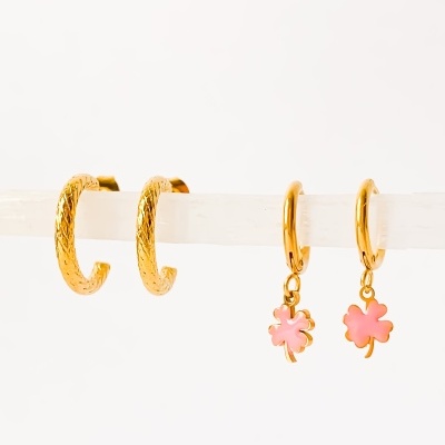 Quatro brincos dourados exibidos numa barra branca, incluindo dois com textura gravada e dois com pendentes em forma de trevo rosa.