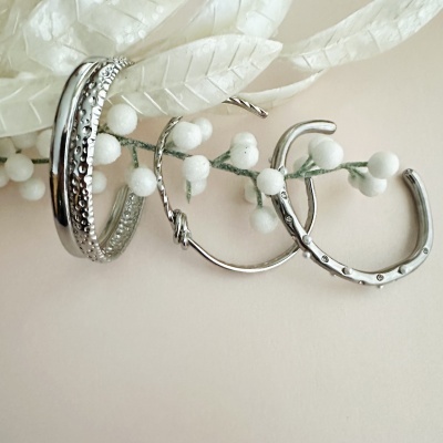 Três braceletes em metal prateado com diferentes texturas e designs sobre fundo claro e folhas brancas artificiais.