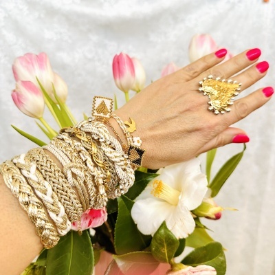 Braço com pulseiras douradas e brancas e anel dourado com detalhes, flores de fundo.