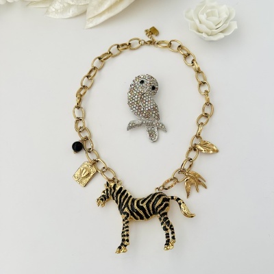 Colar dourado com pendentes de zebra, folha e pássaro, broche de papagaio com pedras, sobre fundo branco com flores.