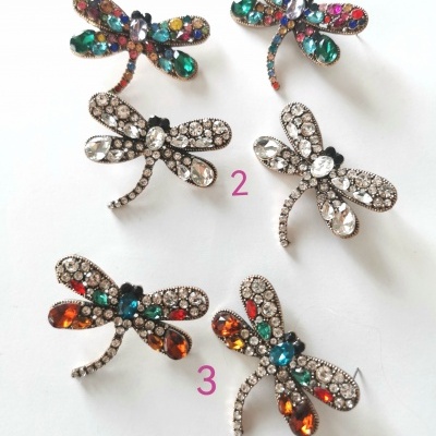 Seis broches em forma de libélula com pedras brilhantes coloridas sobre superfície branca