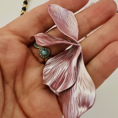 Colar com contas pretas e douradas e pendente rosa em forma de pétalas segurado na mão.