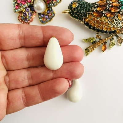 Mão segurando brinco gota branco com broches decorativos coloridos ao fundo