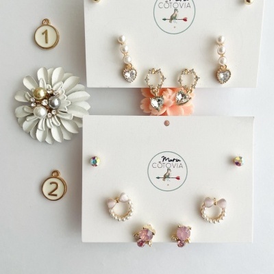 Dois cartões com brincos variados dourados, pérolas e pedras, logótipo Maria COTOVIA e marcadores numerados sobre fundo branco