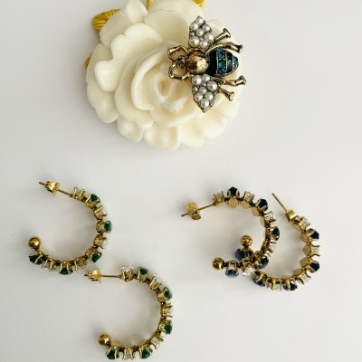 Anel flor branca com abelha dourada e pérolas e dois pares de brincos em argola com pedras verdes e azuis sobre fundo branco