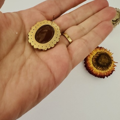 Broche dourado com centro castanho segurado na mão com fundo branco e flores secas ao lado