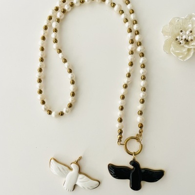 Colar com contas brancas e douradas e pendente de pássaro preto, ao lado pendente de pássaro branco e flor decorativa