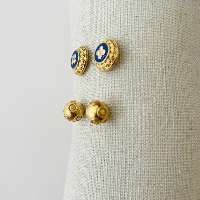 Dois pares de brincos dourados com detalhes em azul e branco sobre tecido bege.