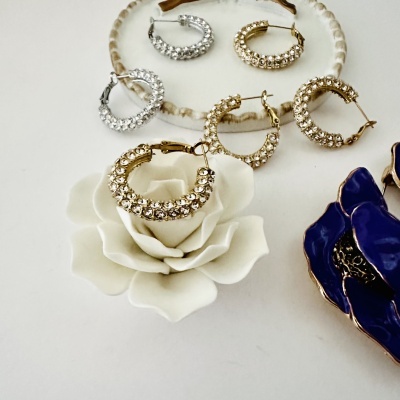 Brincos de argola dourados e prateados com pedras brilhantes em mesa branca com flores decorativas