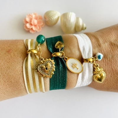 Três pulseiras de tecido com pendentes dourados e contas, usadas no pulso com conchas e flor decorativa ao fundo