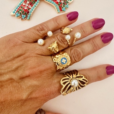 Mão feminina com anéis dourados e pérolas falsas exibidos, fundo claro com decoração colorida