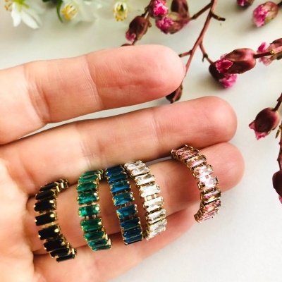 Quatro anéis coloridos com pedras e moldura dourada na mão, com flores em fundo.