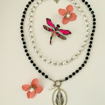 Colares com contas negras e pérolas, brincos florais rosa e broche libélula colorido sobre fundo branco