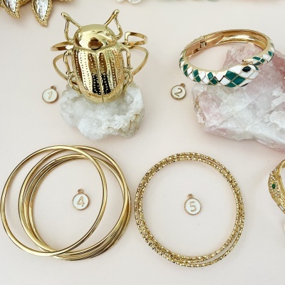 Pulseiras douradas numeradas sobre pedras brancas e rosadas, incluindo escaravelho e padrão xadrez.