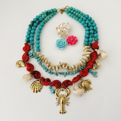 Colar colorido com pérolas azuis, coral vermelho, conchas, e ornamentos dourados sobre fundo branco.