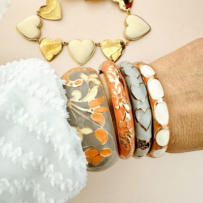 Pulseiras coloridas em cinza, laranja, branco e dourado no pulso com camisa branca e colar de corações ao fundo.