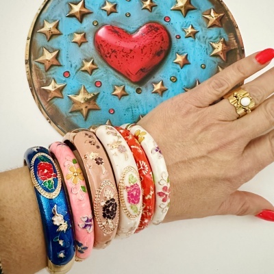 Pulseiras esmaltadas coloridas com motivos florais e detalhes dourados num braço com anel dourado e unhas vermelhas, fundo decorativo azul com coração e estrelas douradas