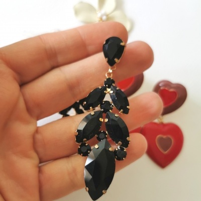 Brinco com pedras pretas facetadas moldura dourada segurado por mão, fundo branco com brincos vermelhos em forma de coração e flor.