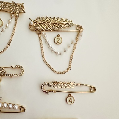 Três alfinetes dourados com pedras e pérolas, com números 2 e 4 em círculos pendurados.