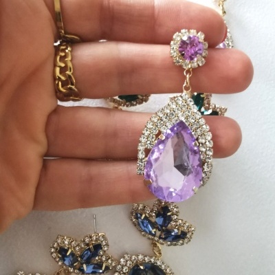 Brinco pendente com pedra roxa em formato de gota e strass em metal dourado segurado por mão