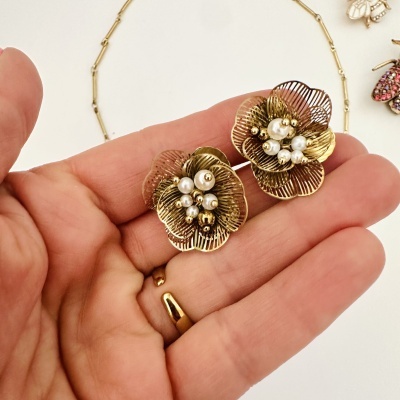 Brincos flor dourada com pérolas em mão, colar dourado e acessórios coloridos ao fundo