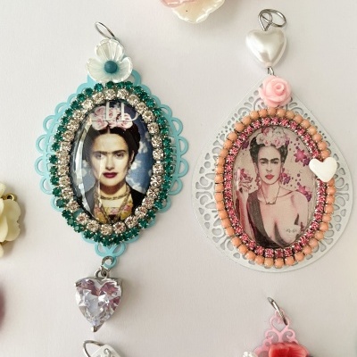 Dois pendentes ovais decorativos com imagens de Frida Kahlo, molduras coloridas com pedras e detalhes florais e corações.