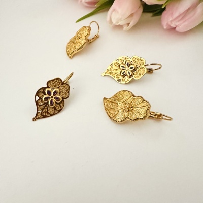 Quatro brincos dourados com design floral e de folhas, dois com detalhes azul-escuros, sobre fundo branco com tulipas cor-de-rosa ao fundo.
