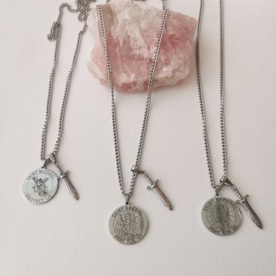 Três colares prateados com pendentes redondos e espadas, sobre fundo branco com pedra rosa.