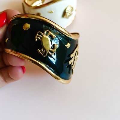 Pulseira esmaltada verde-escuro e branca com detalhes dourados, incluindo caranguejo em relevo, com fundo desfocado