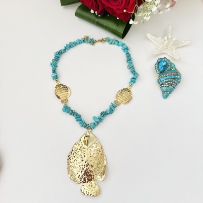 Colar turquesa com pendentes dourados de peixe num ambiente com concha azul, estrela-do-mar branca e flores vermelhas.