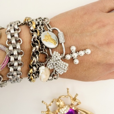 Pulseiras metálicas prateadas e douradas com detalhes de pérolas no pulso, com broche de inseto dourado e roxo ao fundo