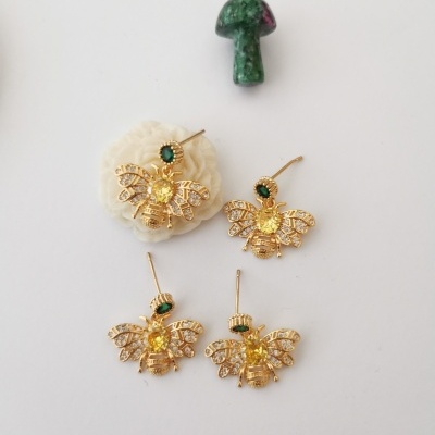 Brincos dourados em forma de abelha com pedras verdes e amarelas e um cogumelo verde e vermelho