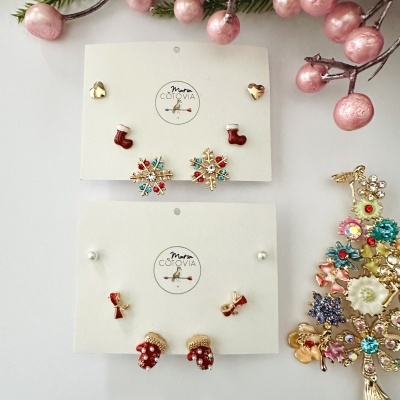 Dois conjuntos de brincos de Natal em cartões brancos com nome Maria Cortiva e enfeites natalícios coloridos ao redor.