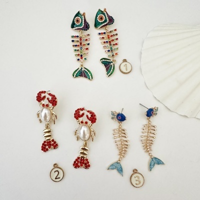 Três pares de brincos coloridos com design de peixes e lagosta sobre fundo branco com concha