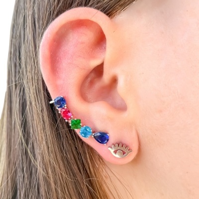 Orelha com dois brincos, pedras coloridas e peça metálica em forma de olho.