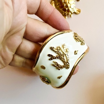 Pulseira branca rígida com detalhes dourados, segurada por uma mão