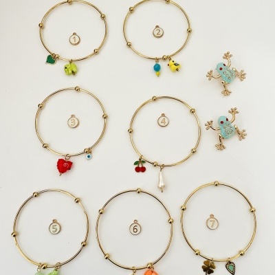 Sete pulseiras douradas com pendentes coloridos variados e dois pendentes de sapo em fundo branco.