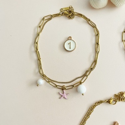 Pulseira dourada com pendentes e pendente número 1 sobre fundo bege com conchas e flor