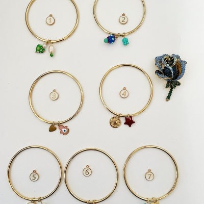 Sete pulseiras douradas com diferentes pendentes coloridos e broche de rosa azul brilhante ao lado.