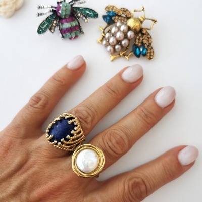 Mão com anéis grandes e broches em forma de abelha com pedras coloridas sobre superfície branca