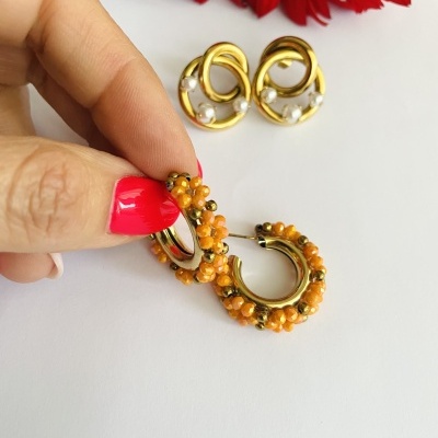 Dois pares de brincos dourados, um com flores laranja decorativas e outro com pérolas, sobre fundo branco