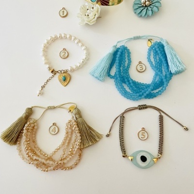 Cinco peças de bijuteria numeradas, incluindo bracelete dourado com azul, pulseira de pérolas branca, pulseira azul com franjas, pulseira bege com correntes e pulseira cinza com olho turco
