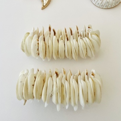 Braceletes feitos de conchas brancas com camadas e broches decorativos ao fundo