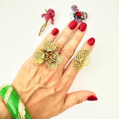 Anéis dourados em design floral e borboleta, pulseira verde com penas douradas, broches de flor e abelha com pedras vermelhas, mão com unhas vermelhas.