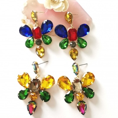 Dois pares de brincos coloridos em formato de borboleta com várias pedras brilhantes sobre fundo branco e uma flor branca ao fundo