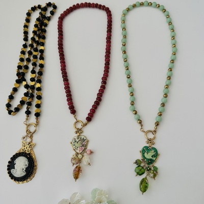 Três colares com contas de cores preto, vermelho e verde com pendentes decorativos em forma de coração e medalhão, dispostos sobre fundo branco.