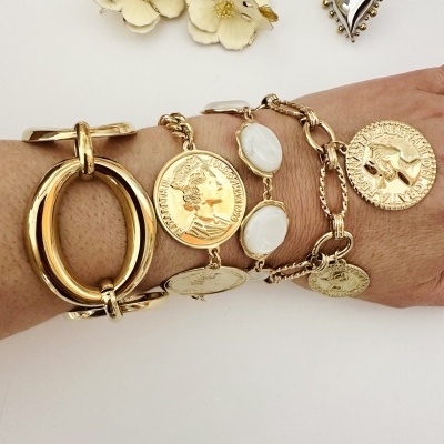 Pulseiras douradas com moedas e pedras brancas num pulso, brincos flor creme e brincos coração prateados ao fundo