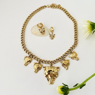 Conjunto de colar e brincos dourados com pendentes de cabeça de animal e cristais em fundo branco com flores