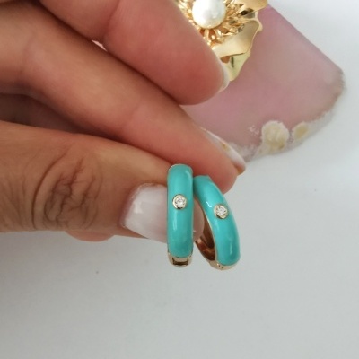 Brincos dourados com esmalte azul turquesa e pedra branca, segurados por mão, fundo com flor dourada.